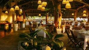 An indoor dining area with multiple tables and chairs, a central planter with greenery, and hanging lamps. - Nosy Be Hotel & Spa (Nosy Be)