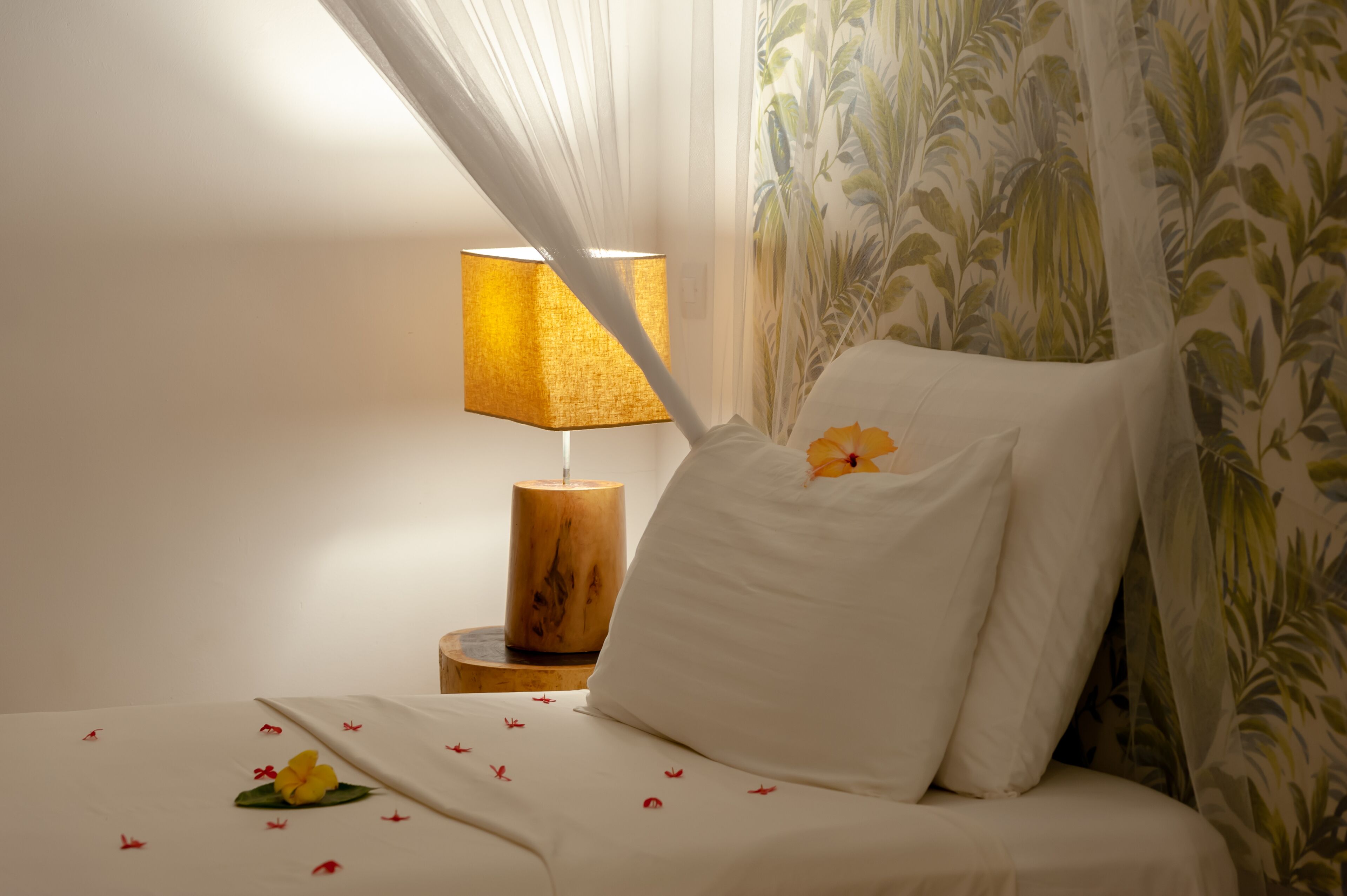 Ein Bett mit weißen Leinen, ein Nachttisch aus Holz mit einer Lampe und ein blumiger Vorhang.