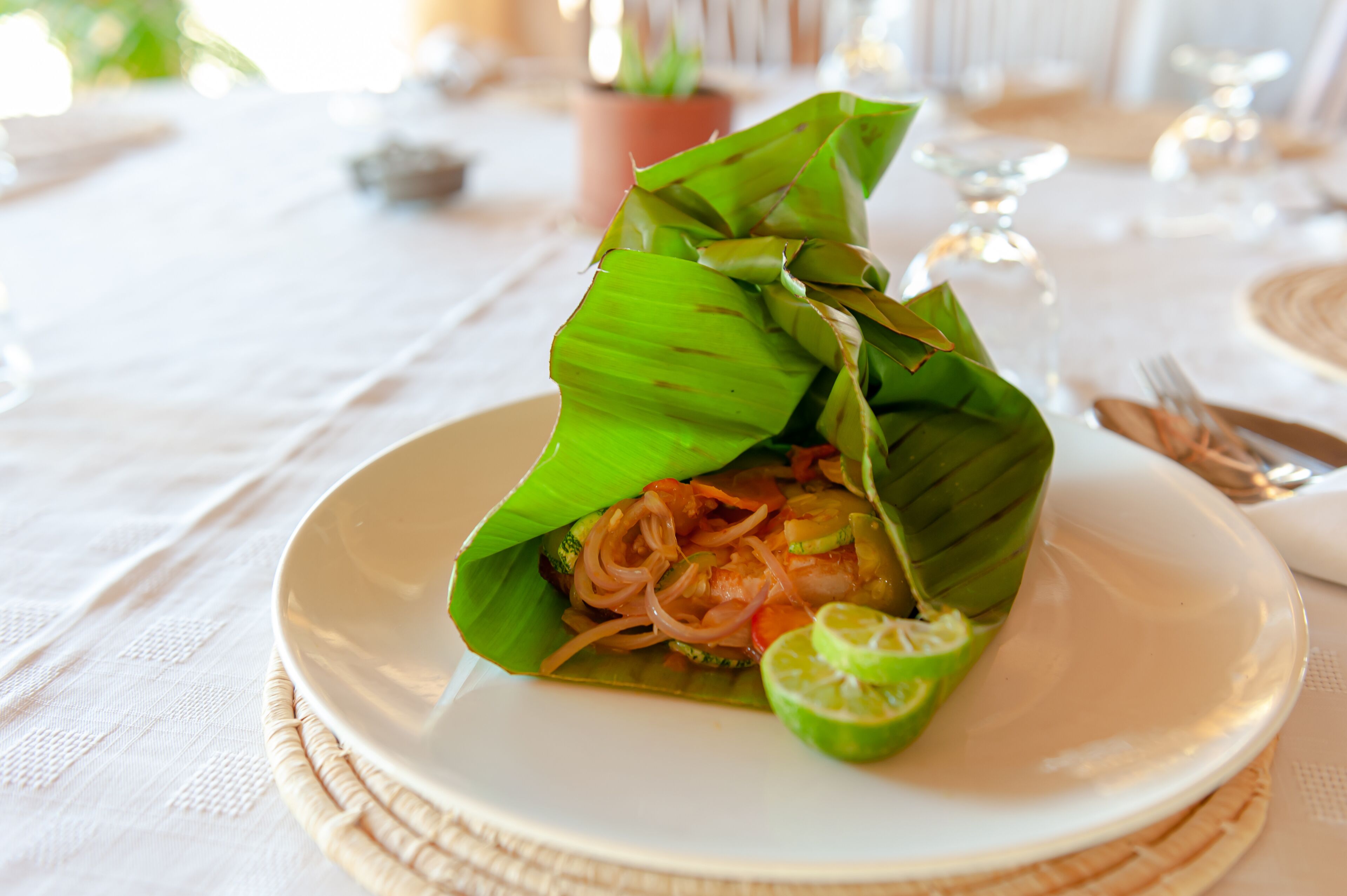 A plate of food wrapped in banana leaves, garnished with lime, on a white tablecloth.