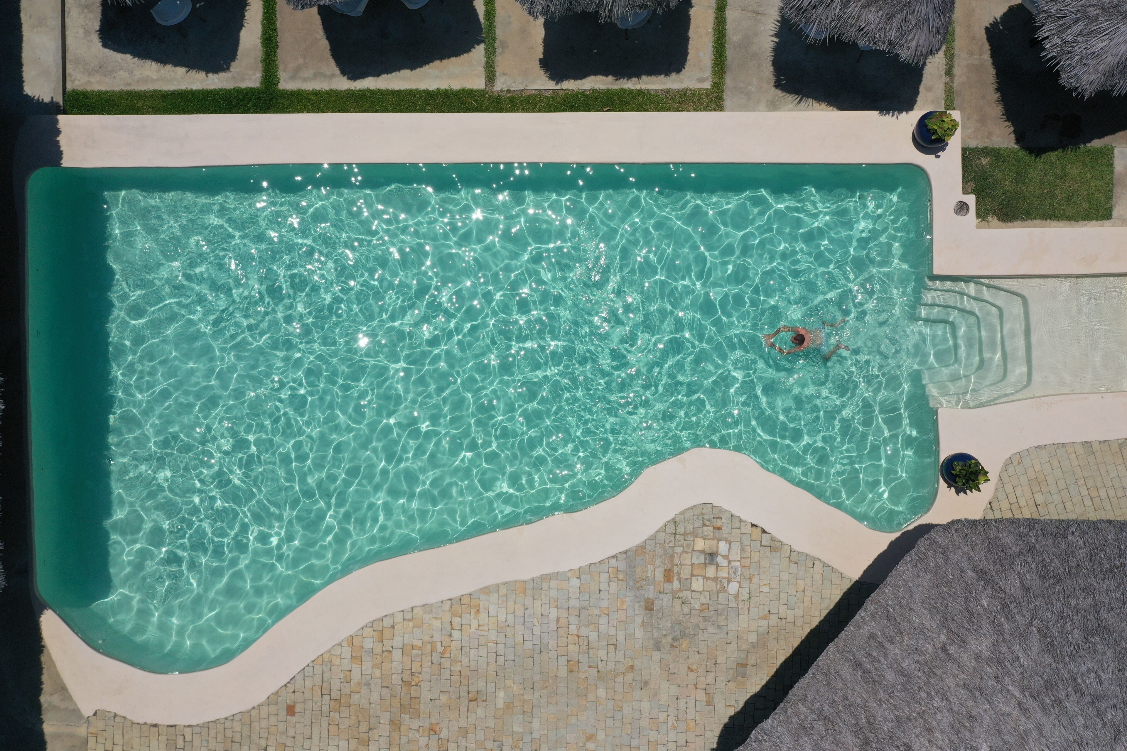 An aerial view of a swimming pool with clear water, surrounded by a tiled deck and grass areas.