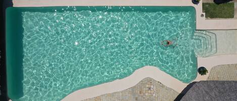 An aerial view of a swimming pool with clear water, surrounded by a tiled deck and grass areas.
