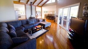 A spacious living room with a large sectional sofa, a fireplace, wooden beams, and a dining area visible through large windows. - The White House (Norfolk Island)