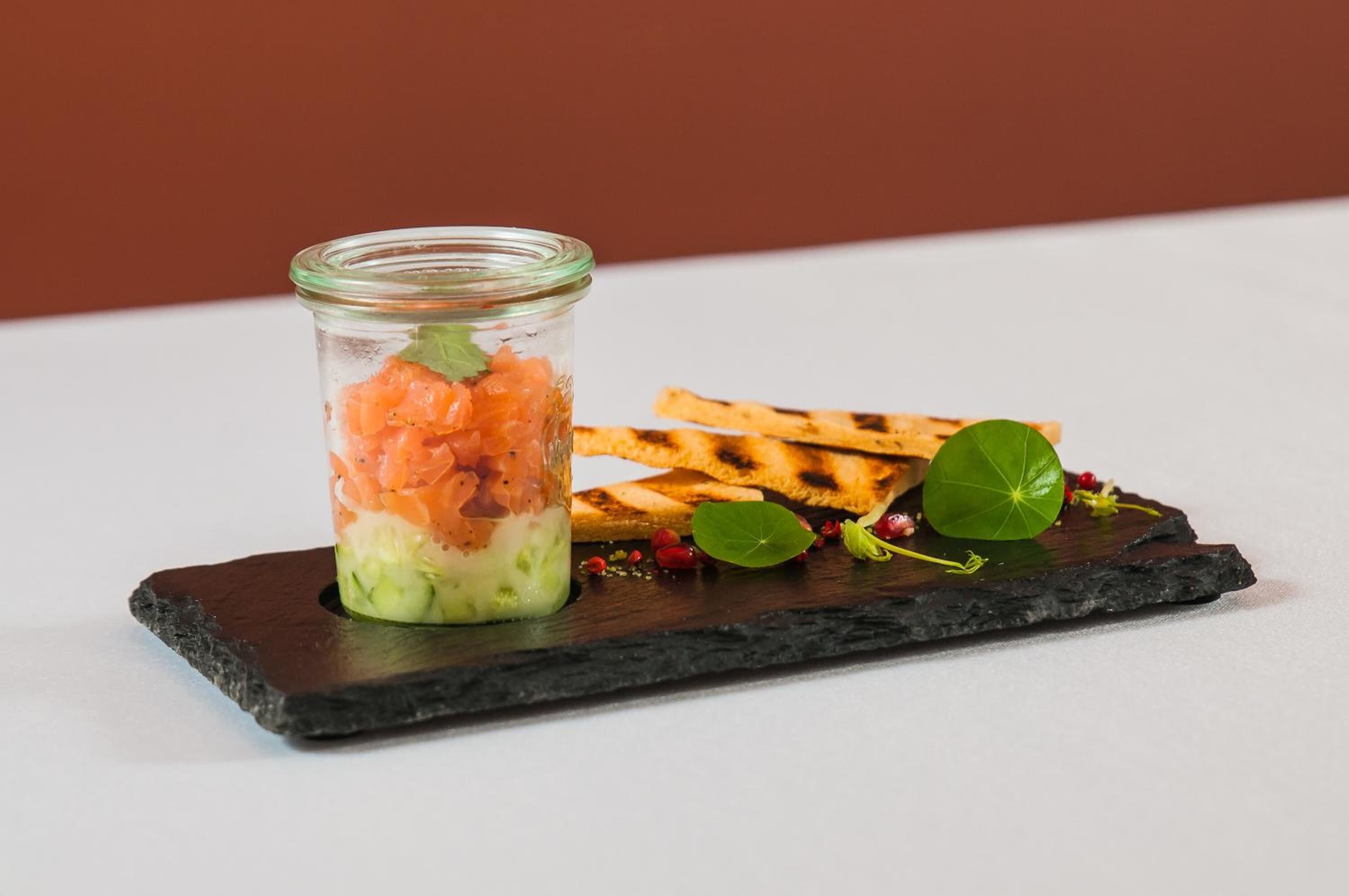 A glass jar with a layered salad, accompanied by grilled bread and garnished with a leaf and berries on a slate board.