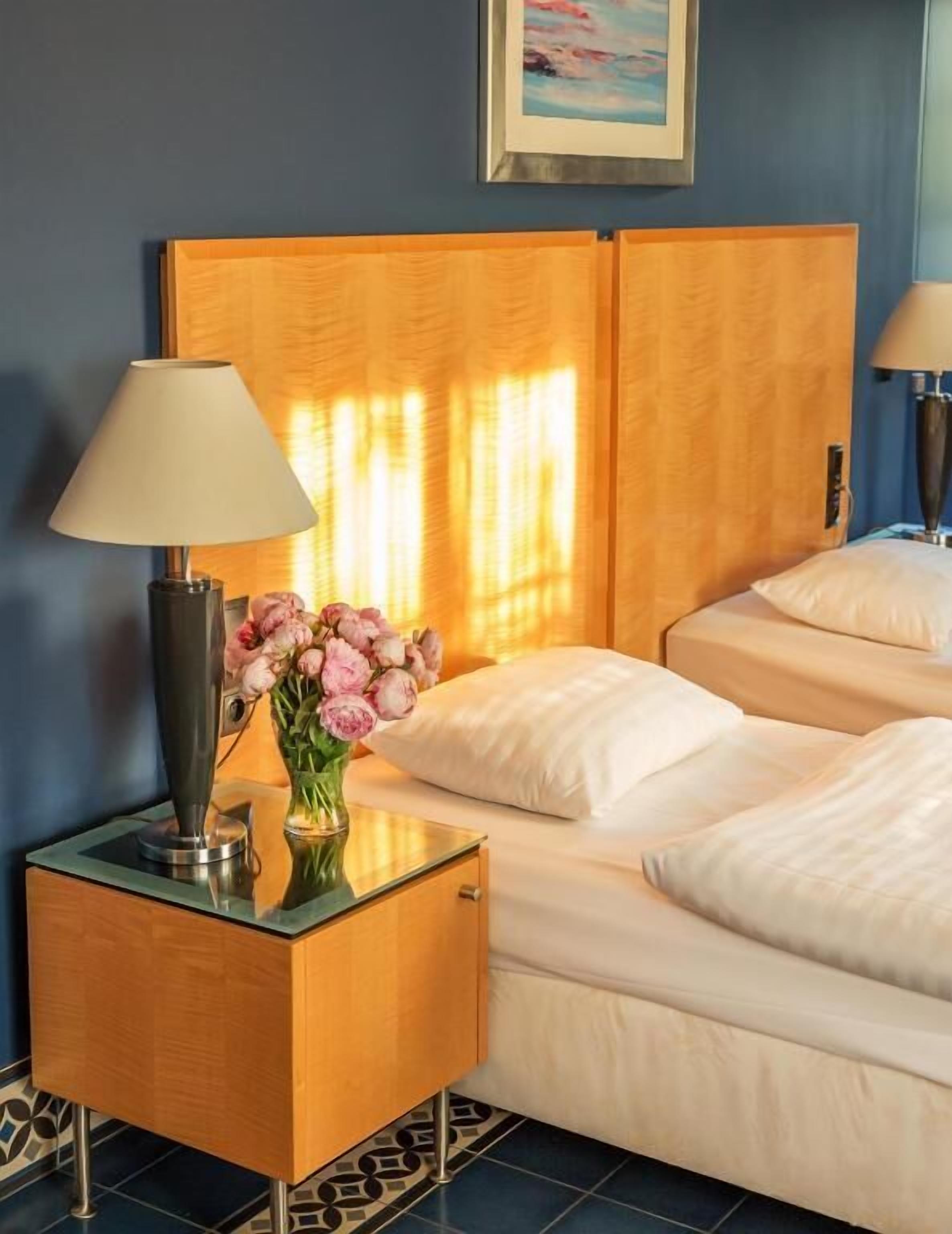 A hotel room with a bed, a bedside table with a lamp, and a vase of flowers.