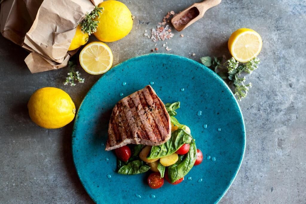 Ein Teller mit gegrilltem Fleisch und Salat, Zitronen und einer braunen Papiertüte auf einer strukturierten Oberfläche.