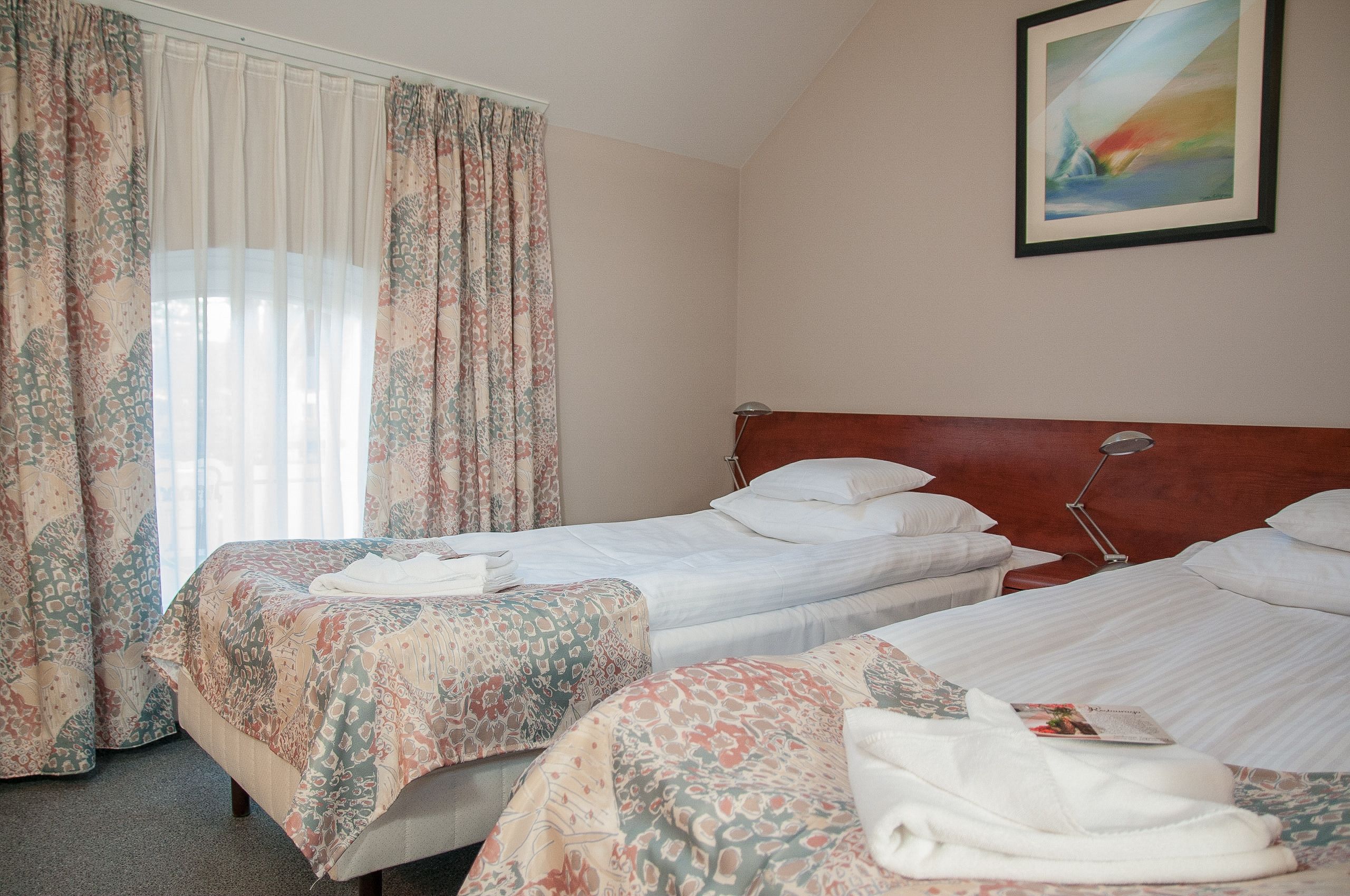 Ein Hotelzimmer mit zwei Betten, floralen Vorhängen, einem Bild an der Wand und einer Nachttischlampe.