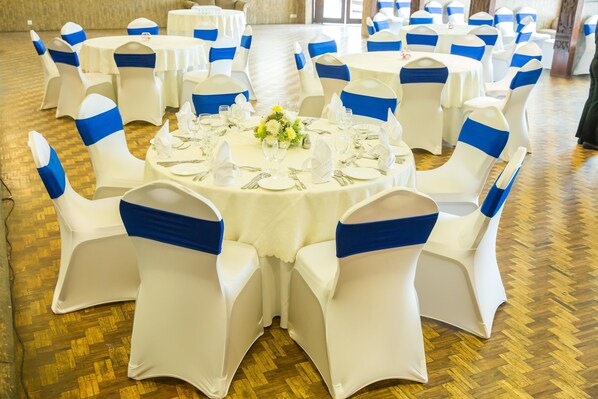 Un salón de banquetes con mesas redondas dispuestas para un evento formal, cada una con manteles blancos y cintas azules en las sillas.