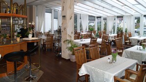 Un restaurante con mesas y sillas de madera, una zona de bar con taburetes y amplias ventanas que dejan entrar luz natural.