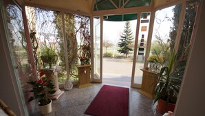 Entrada de hotel con alfombra roja, plantas en macetas y vistas al exterior.