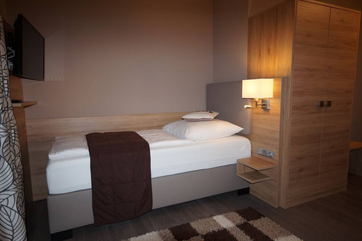 Habitació d'hotel moderna amb un llit individual, una taula de nit, una làmpada i un armari.