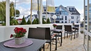 Une terrasse extérieure avec des chaises et des tables en osier, un pot de fleurs aux fleurs rouges, et une vue sur des bâtiments modernes en arrière-plan.