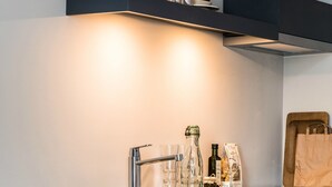 A modern kitchen with a dark countertop, a wooden stool, and a shelf with wine glasses and bottles. - Bold Hotel München Giesing (Munich)