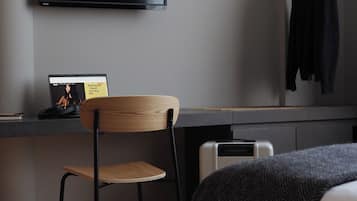 A modern hotel room with a flat-screen TV mounted on the wall, a wooden chair, a small desk with a laptop, and a bed with a dark throw blanket.