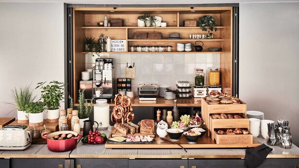 A kitchen with wooden shelves, a coffee machine, and various food items. - Bold Hotel München Giesing (Munich)