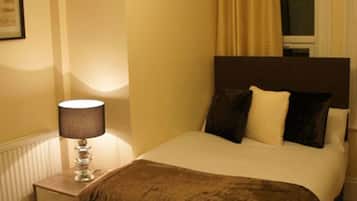 Une chambre à coucher avec un lit, une table de chevet avec une lampe, une fenêtre avec des rideaux et un radiateur.