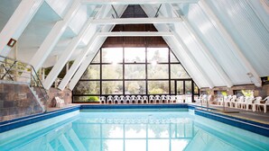 An indoor swimming pool with a clear view of the outdoors through large windows, surrounded by white chairs. - Termas de Aguas Calientes (Puyehue)