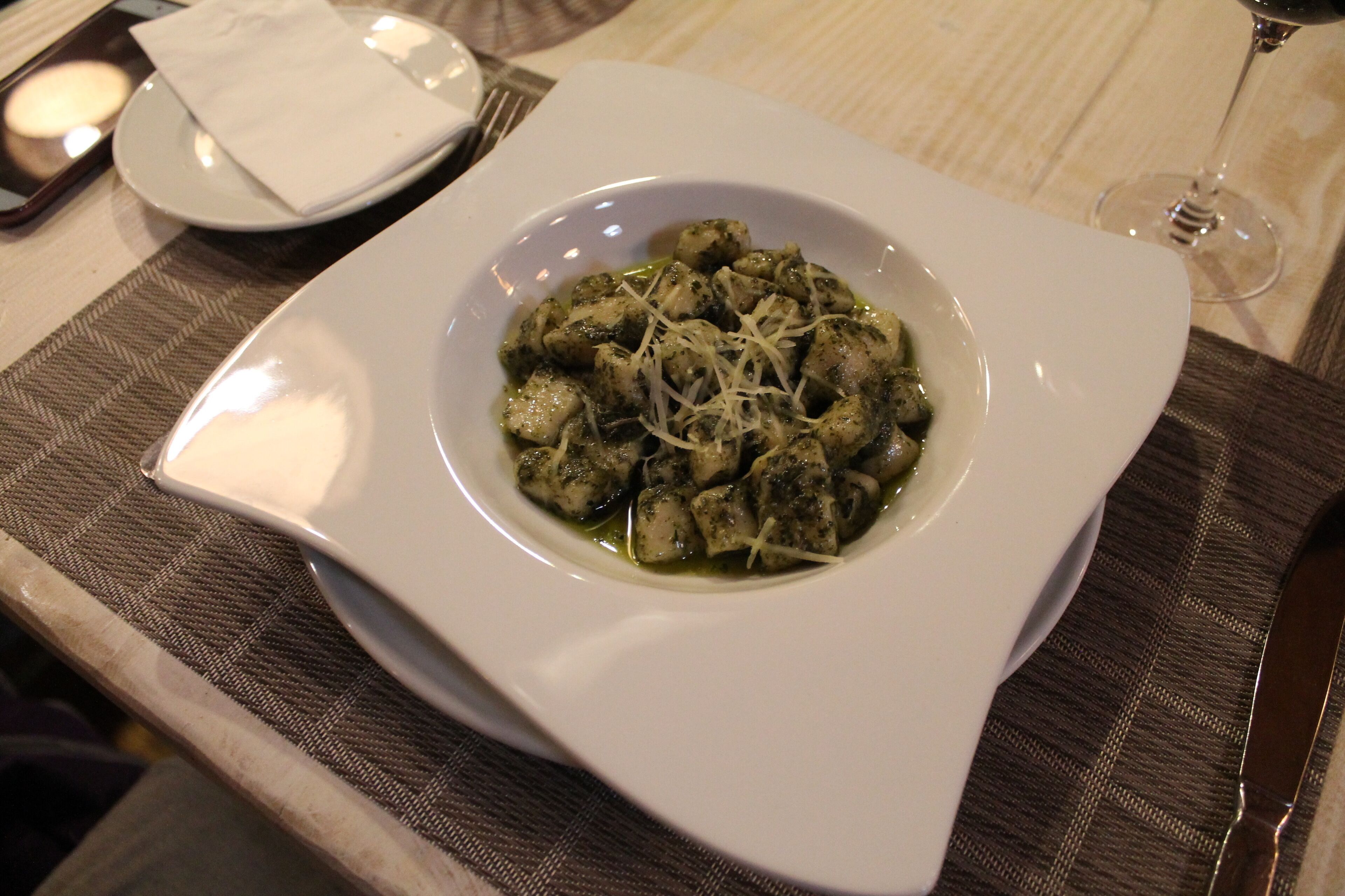 A square white plate with a dish of food, possibly gnocchi, garnished with grated cheese, on a table with a placemat.