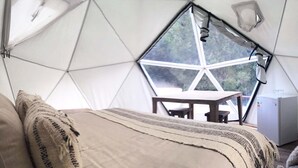 A cozy interior of a geodesic dome with a bed, a table, and a view of the outdoors through a large window. - Termas de Aguas Calientes (Puyehue)