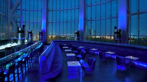 A modern bar with a panoramic city view, featuring a long counter, seating area, and ambient lighting.