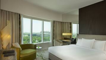 Habitación de hotel con una cama grande, un sillón verde, una mesita de noche con lámpara y vistas a la ciudad a través de amplios ventanales.