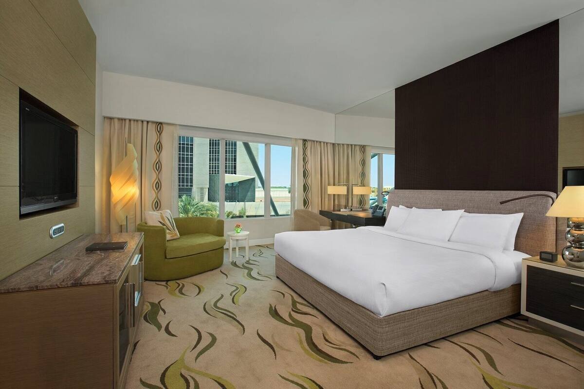 Quarto de hotel moderno com uma cama grande, uma mesa de escritório, uma televisão e vista para um edifício.