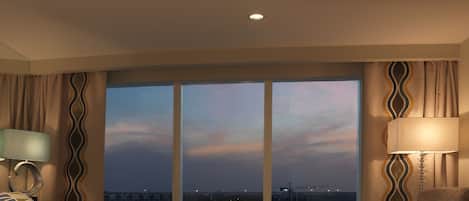 A hotel room with a large window, a bed, a chair, and a side table with a lamp.