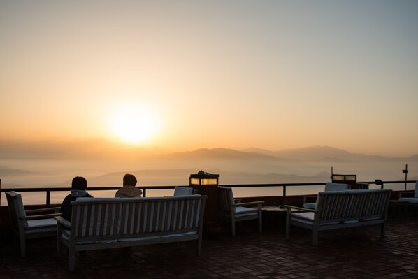 Zonsondergang vanaf een terras met houten banken en tafels, met uitzicht op een waterpartij en verre bergen.