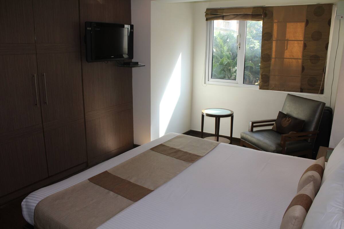 Ein Hotelzimmer mit einem Bett, einem an der Wand befestigten Fernseher, einem Fenster mit Vorhängen und einem Sessel.