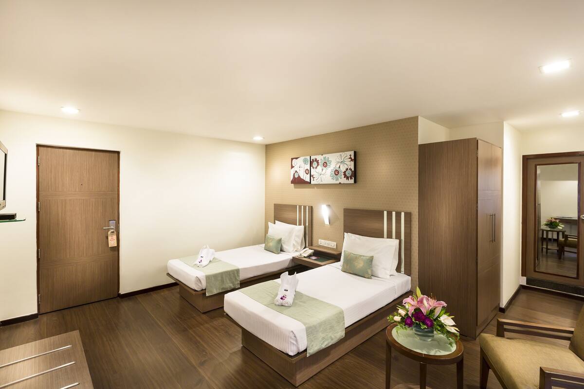 Ein Hotelzimmer mit zwei Betten, einem kleinen Tisch mit Blumen in einer Vase, einem Spiegel und einem Holzschrank.