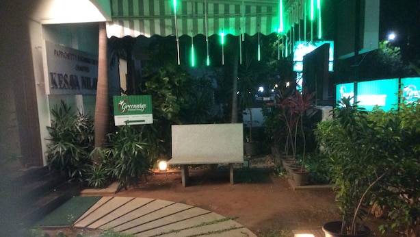 Een buitenruimte bij nacht met een bordje voor 'Greenings', een bank, potplanten en een pad dat naar een gebouw leidt.