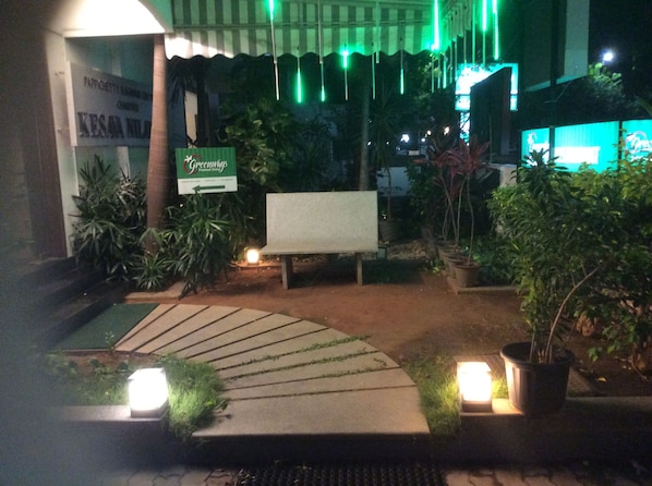 Ein Außenbereich bei Nacht mit einem Schild für 'Greenings', einer Bank, Topfpflanzen und einem Weg zum Eingang eines Gebäudes.