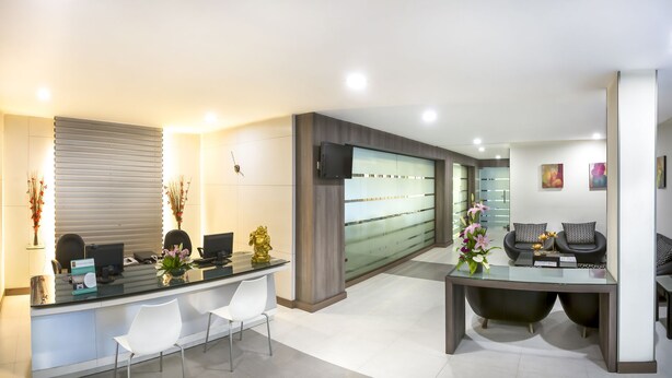 A modern reception area with a glass counter, white chairs, a TV, and a large window with blinds. - The Lotus - Apartment Hotel, Venkatraman Street (Chennai)