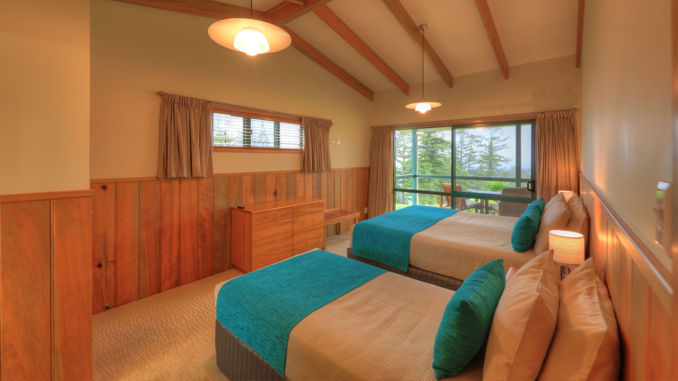 A bedroom with two beds, wooden paneling, a dresser, and large windows with curtains.