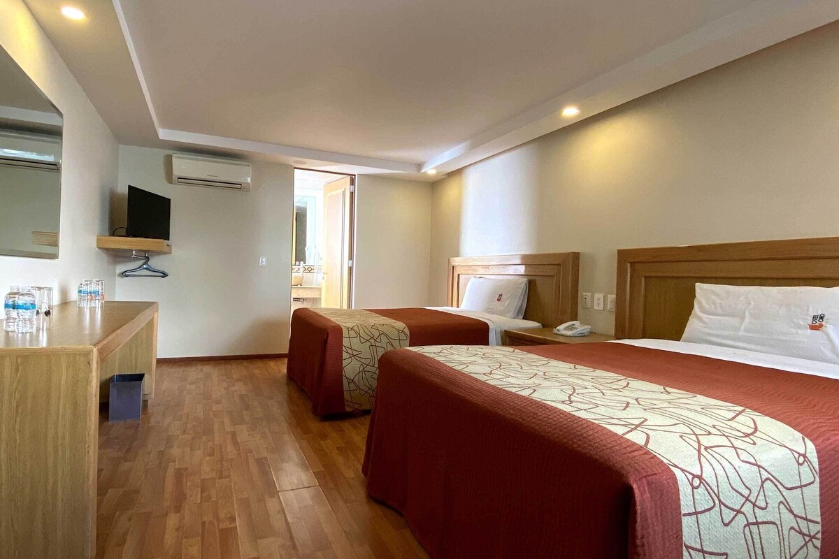 A hotel room with a bed, bedside tables, a desk, a mirror, and an air conditioning unit.