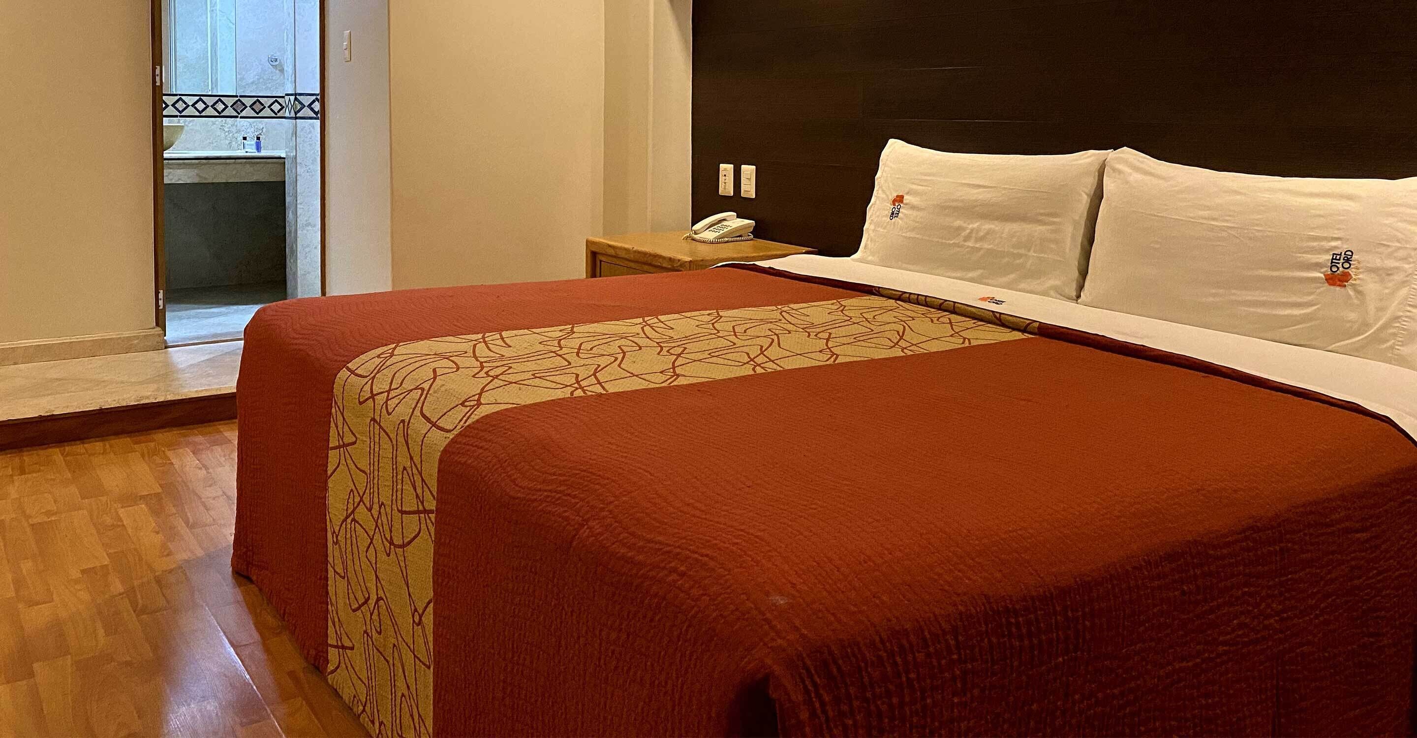 Quarto de hotel com uma cama grande, uma mesinha de cabeceira com telefone e vista para um banheiro com pia e espelho.