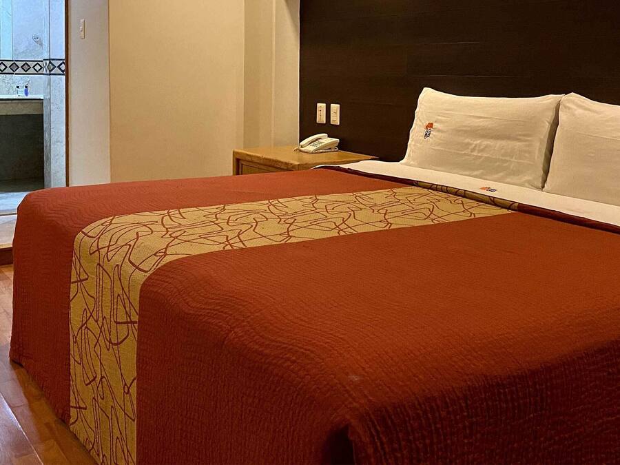 Quarto de hotel com uma cama grande, uma mesinha de cabeceira com telefone e vista para um banheiro com pia e espelho.