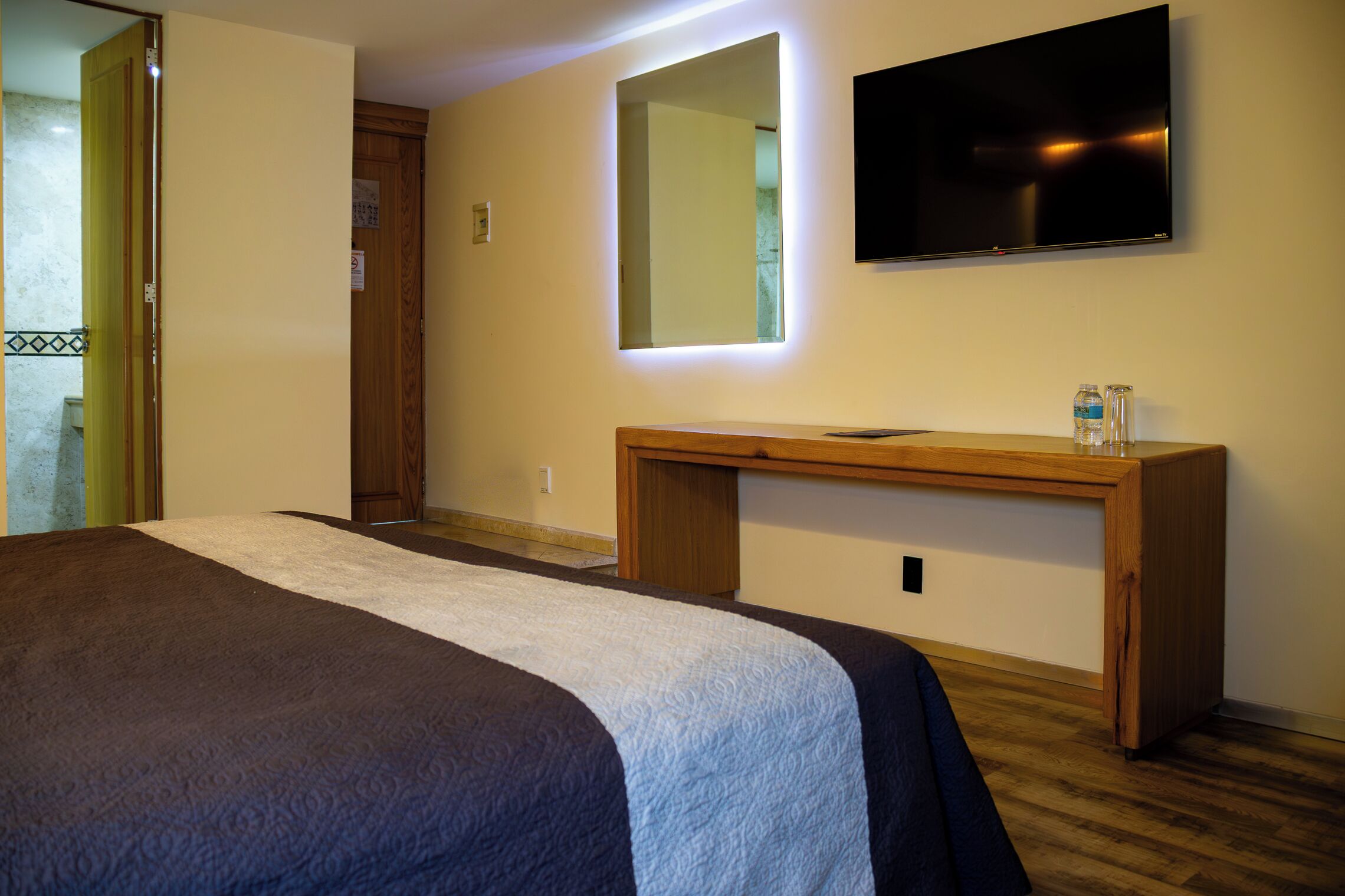 Quarto de hotel com uma cama, uma escrivaninha de madeira, televisão de tela plana e um espelho.