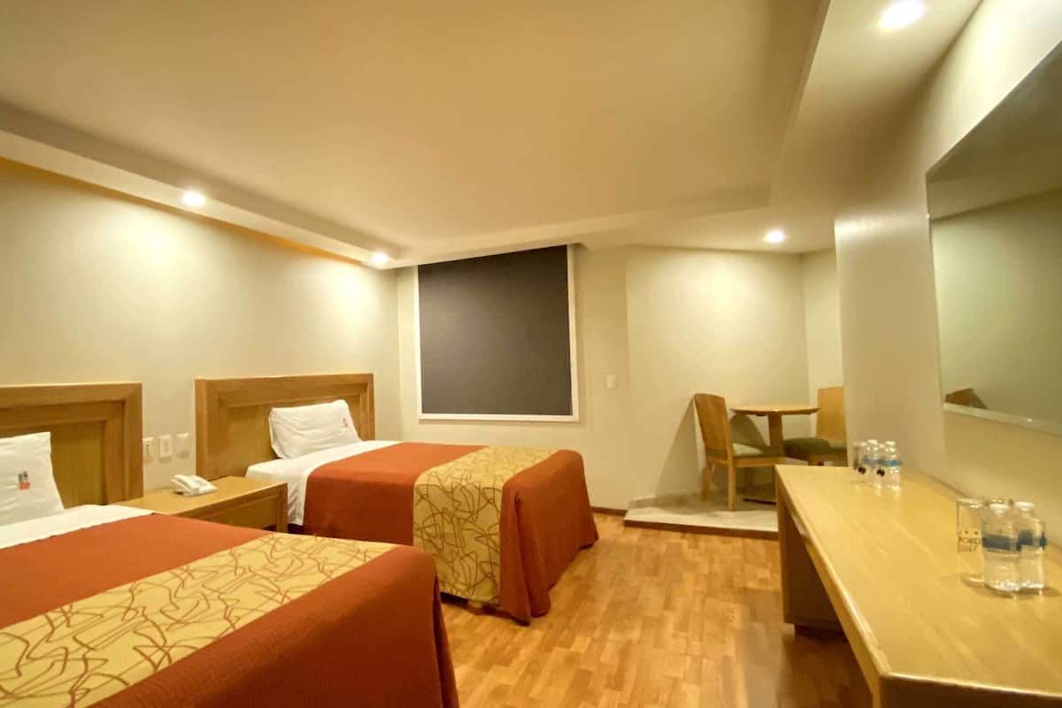 Une chambre d’hôtel avec deux lits, un bureau, une chaise et une petite table sur laquelle se trouvent des bouteilles d’eau.