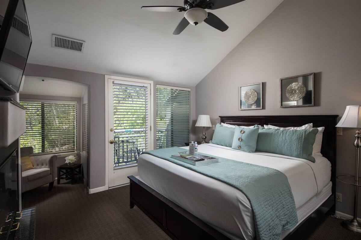 A bedroom with a bed, bedside lamps, a ceiling fan, and two windows with blinds.