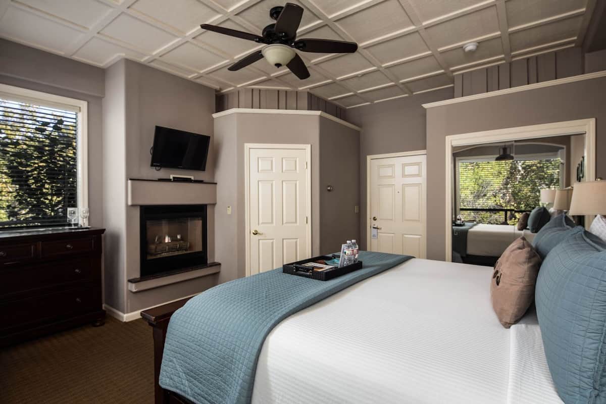 A hotel room with a bed, a TV mounted above a fireplace, a ceiling fan, and a door leading to a balcony.