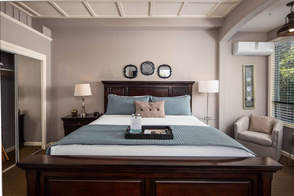 A neatly arranged bedroom with a large bed, two bedside tables, a nightstand, a chair, and a window with blinds.