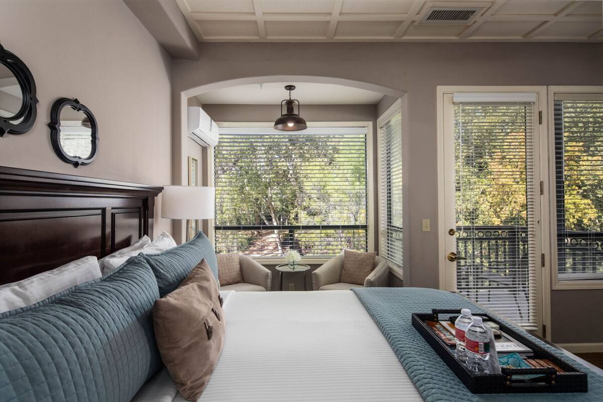 A hotel room with a large bed, a bedside table with a lamp, a small round table, and a view of trees through the window.