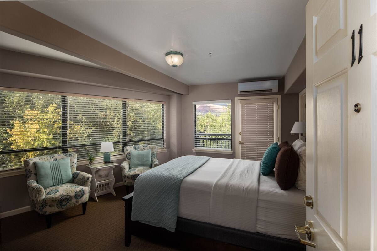 A bedroom with a bed, two chairs, a nightstand, a window with blinds, and an air conditioning unit.