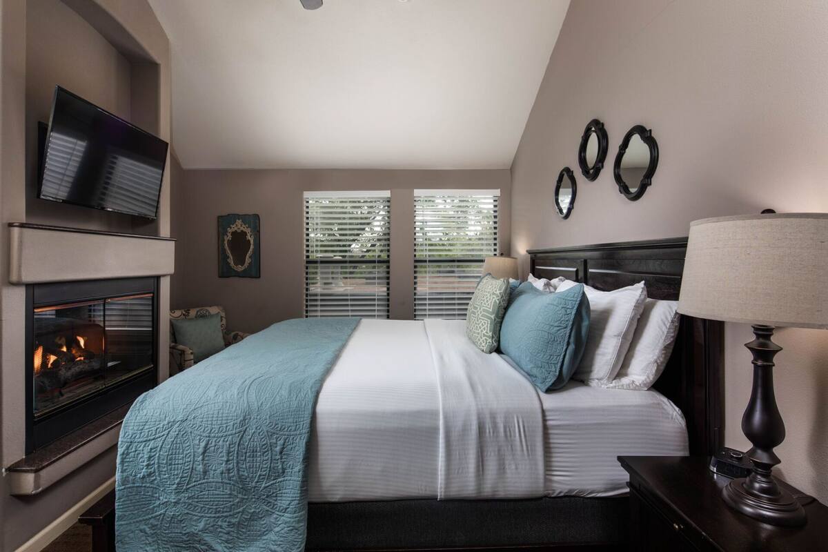 A bedroom with a fireplace, a bed with multiple pillows, a bedside table with a lamp, and a TV mounted on the wall.