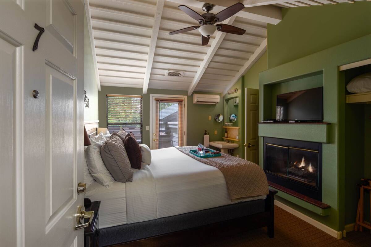 A bedroom with a bed, a ceiling fan, a fireplace, and a television on a shelf.