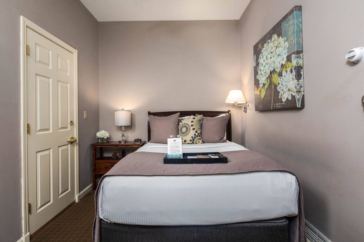 A hotel room with a bed, bedside lamps, a nightstand, a door, and a wall-mounted artwork.