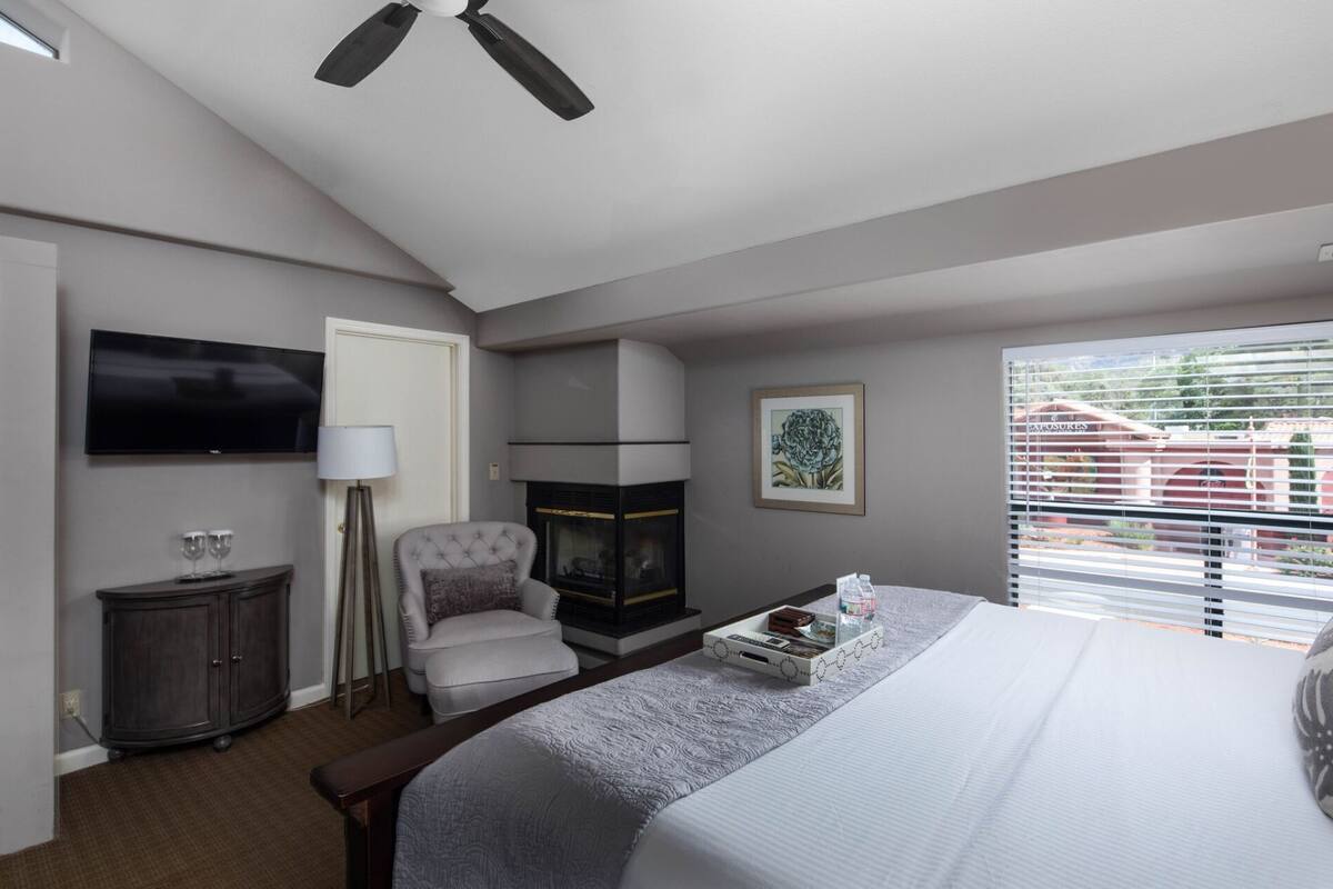 A hotel room with a bed, a television, a fireplace, a chair, and a window with blinds.