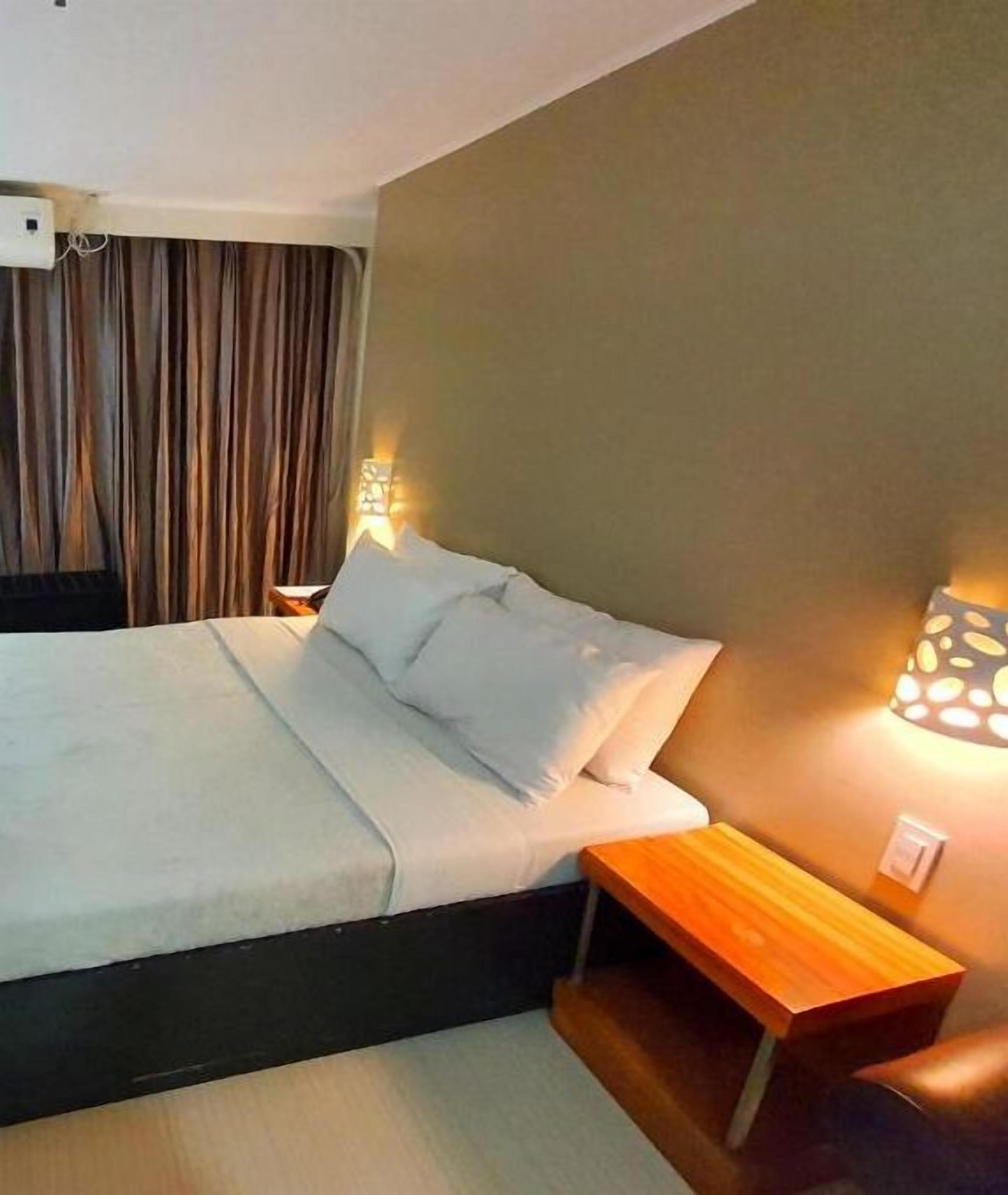 A neatly made bed with white linens, a wooden nightstand with a lamp, and a curtain in the background.