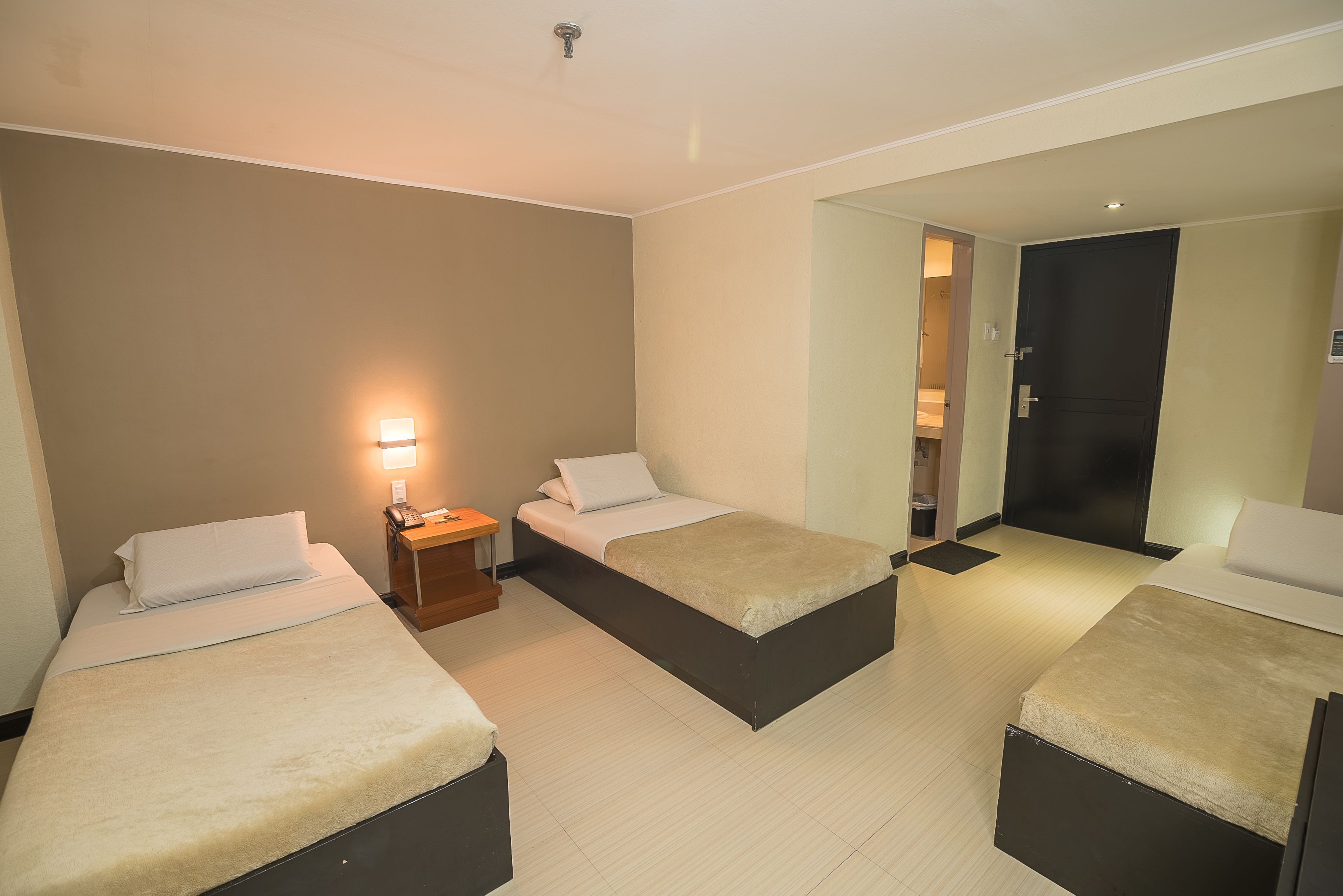 Ein Hotelzimmer mit drei Einzelbetten, einem Nachttisch mit Lampe und einer Tür zu einem Badezimmer.