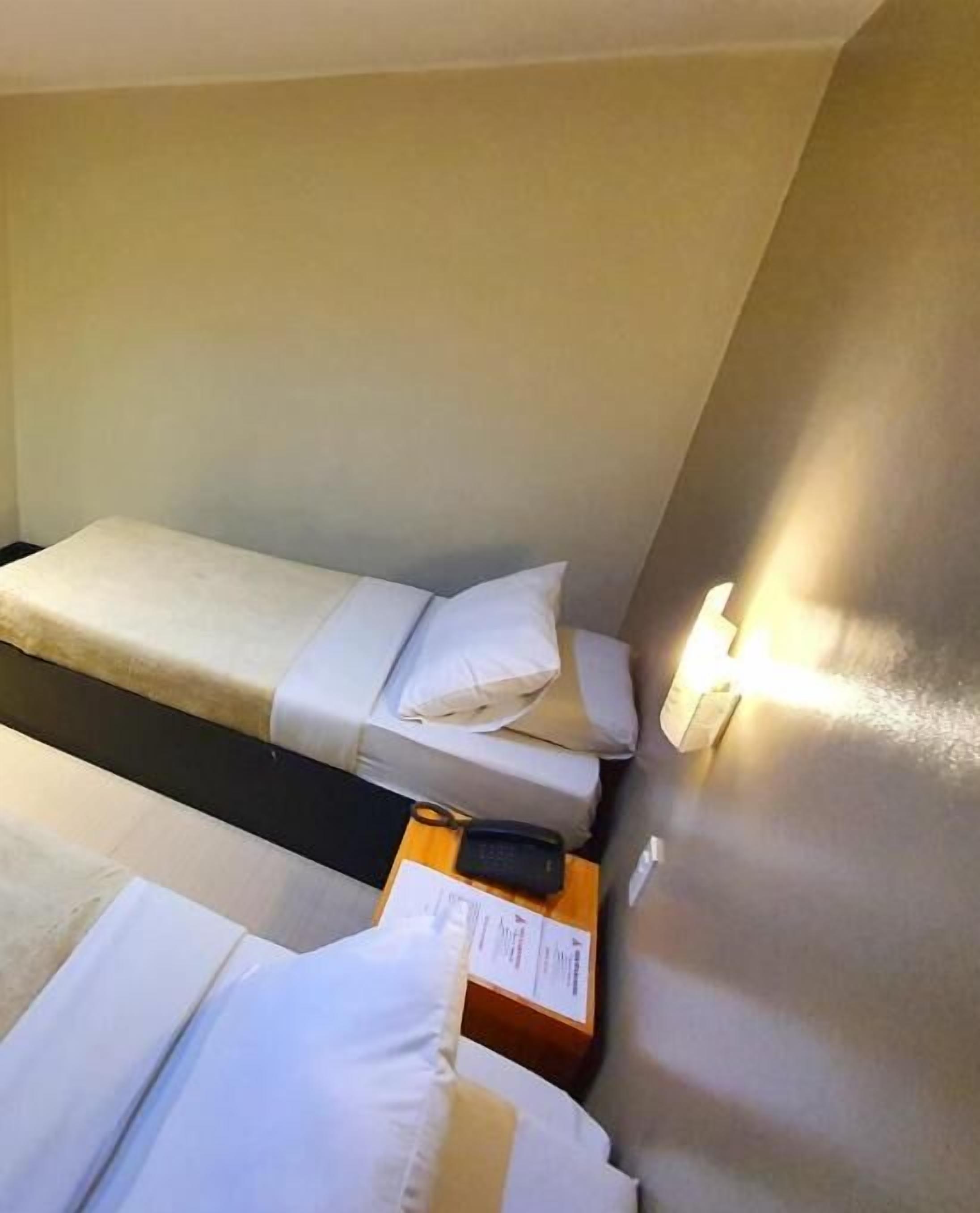 A small, single-bed room with a bedside table, a lamp, and a power outlet.